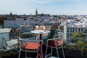 Una terraza en la azotea con dos sillones de mimbre blancos y una mesa, con vistas a un paisaje urbano que incluye edificios y una torre.