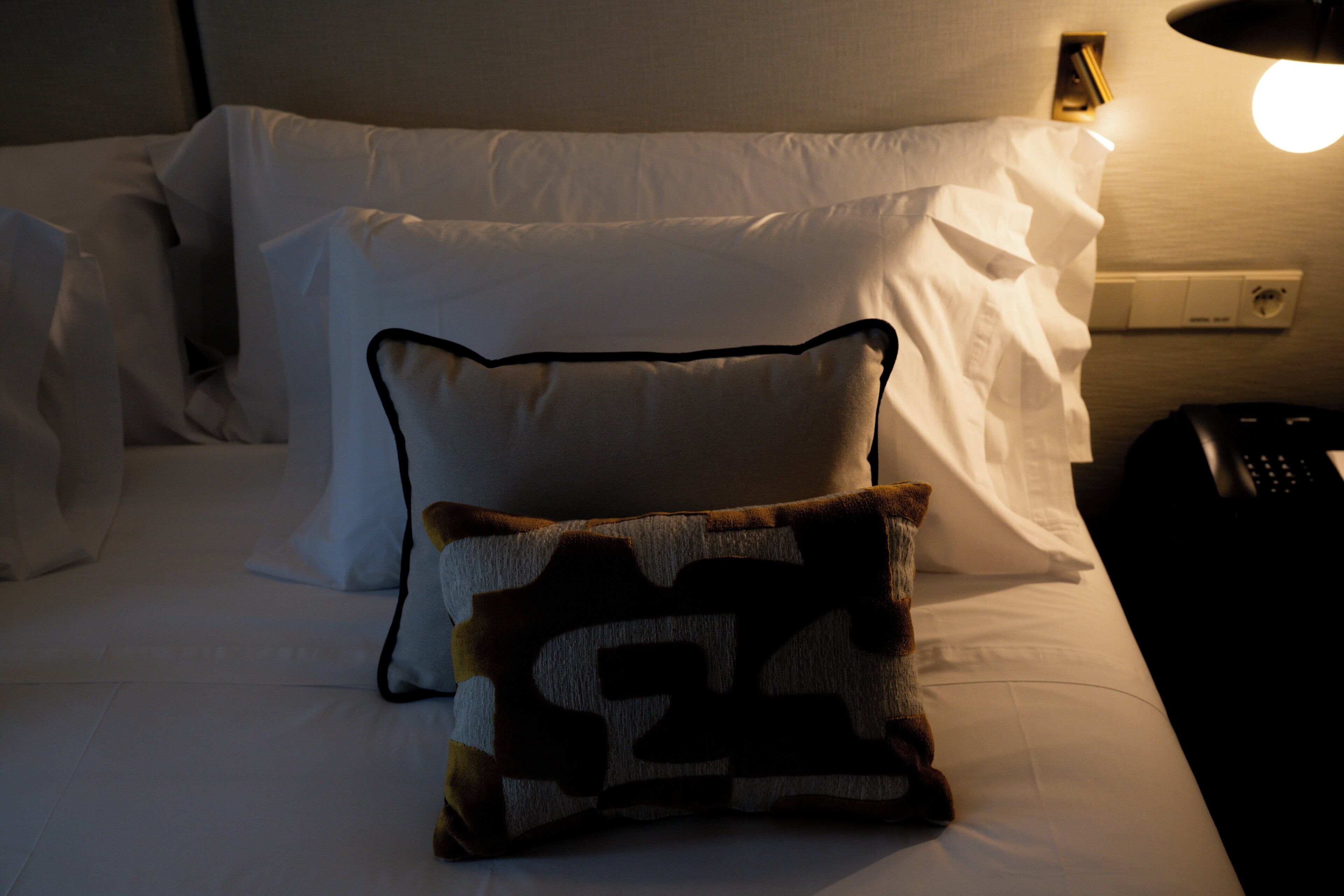Een bed met witte lakens, een kussen met dessin, een afstandsbediening en een stopcontact.