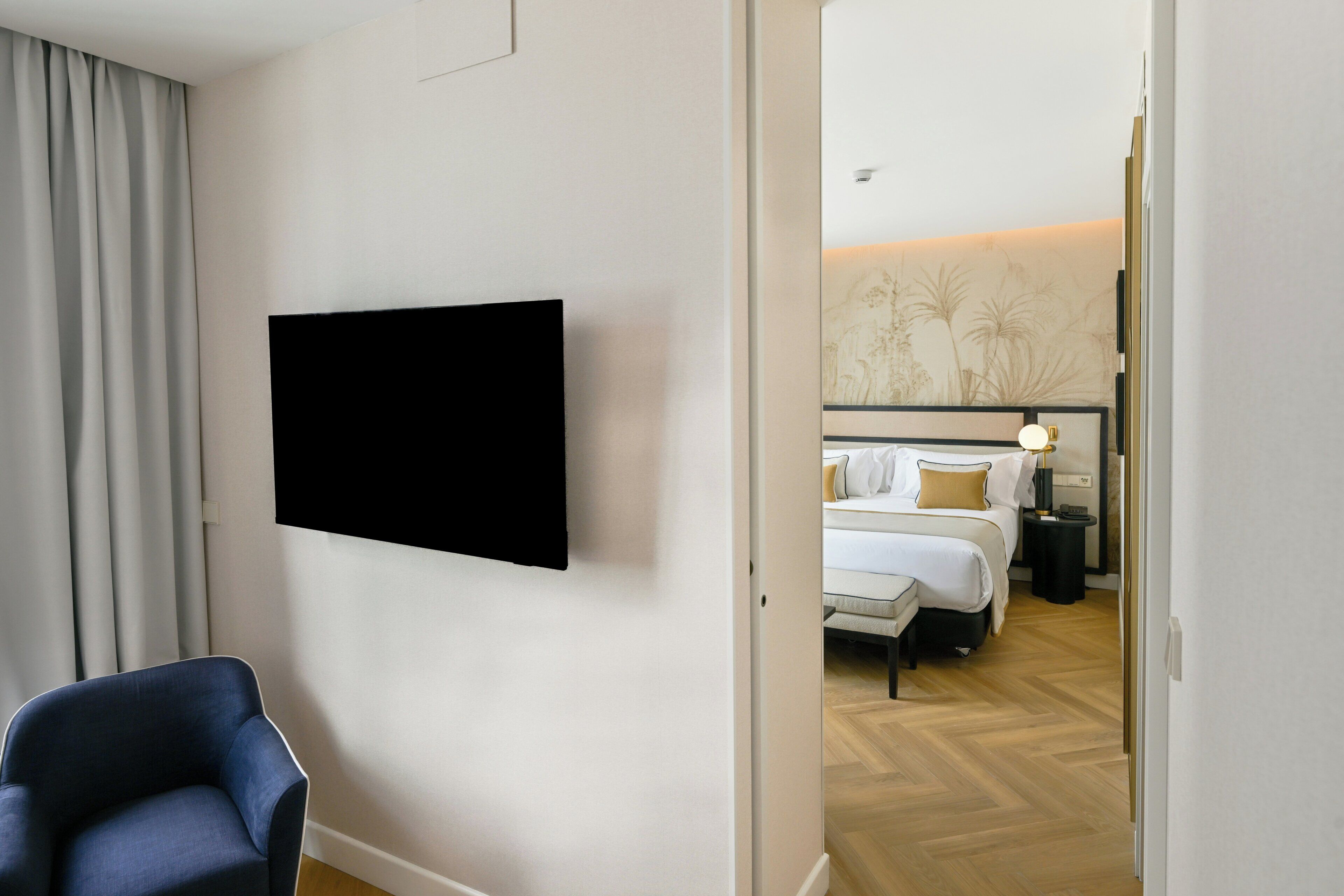 Habitación de hotel con televisor de pantalla plana en la pared, cama con ropa blanca, mesita de noche con lámpara y una silla.