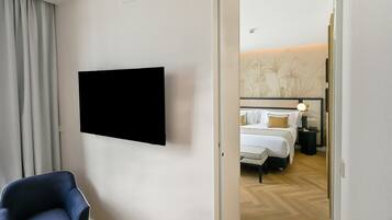 Habitación de hotel con televisor de pantalla plana en la pared, cama con ropa blanca, mesita de noche con lámpara y una silla.