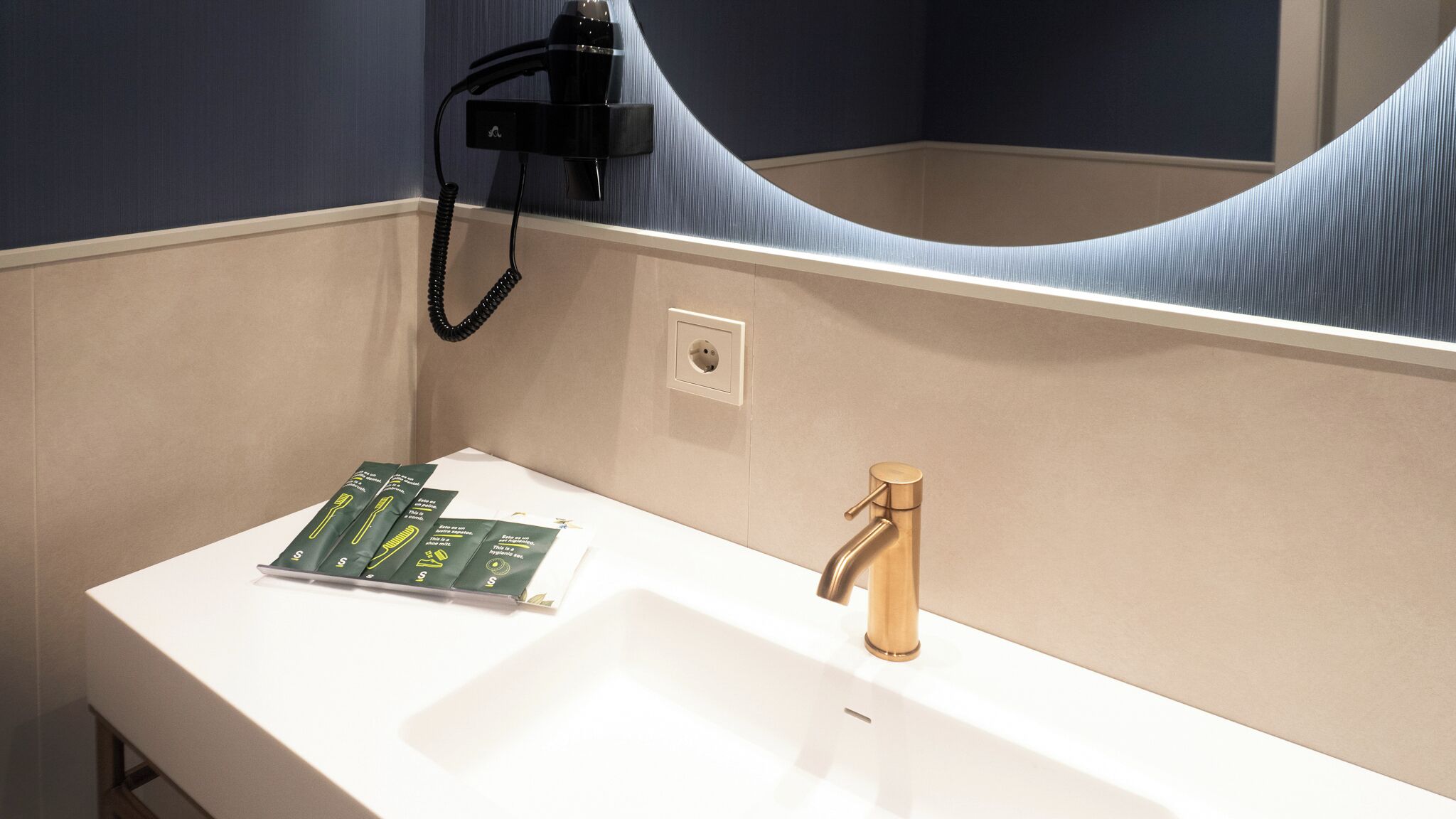 A bathroom with a white sink, a gold faucet, and a mirror reflecting the room. There are green and yellow toothbrushes on the counter.
