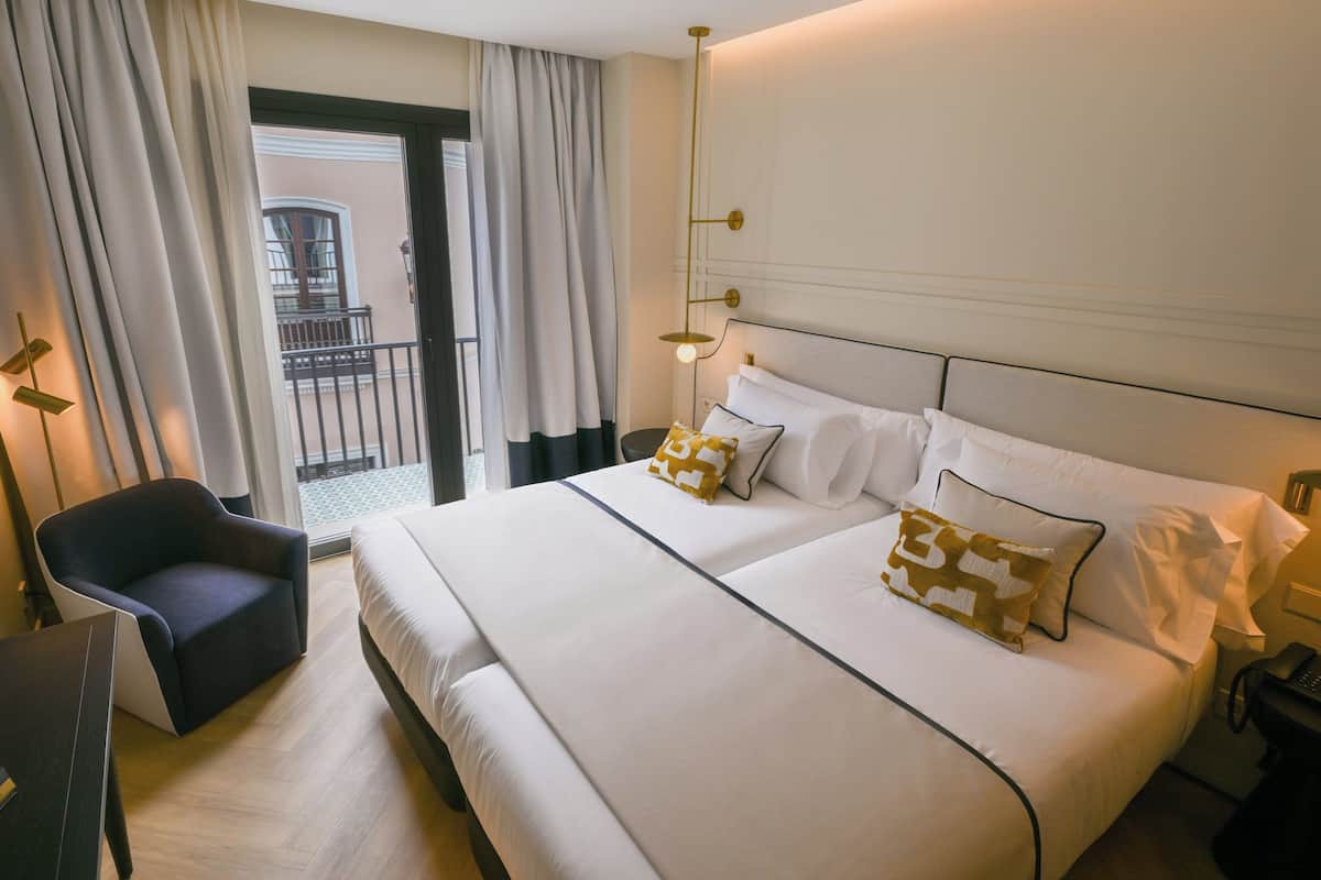 A hotel room with a large bed, two pillows, a chair, a bedside table, and a view of the outside balcony.