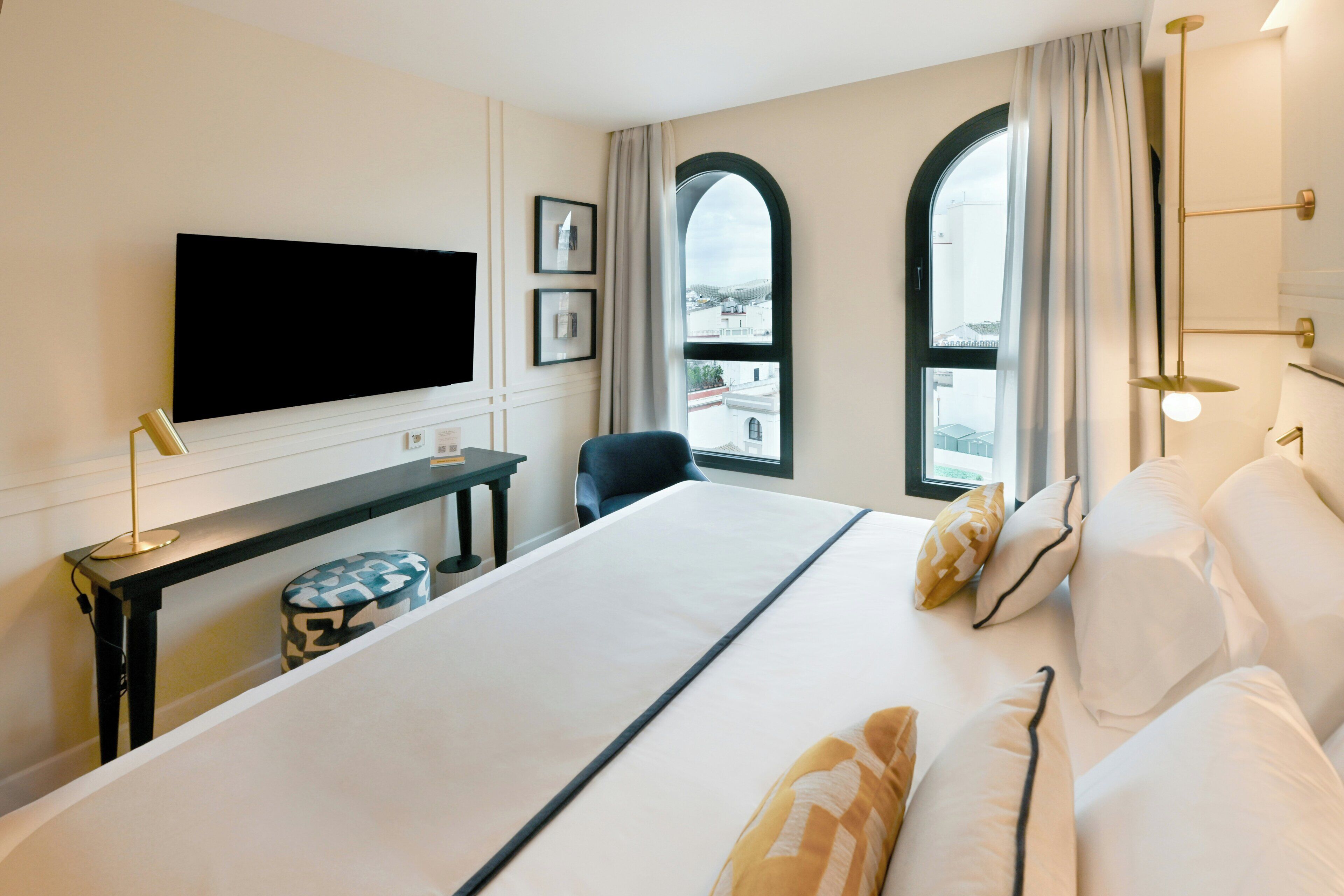 A hotel room with a bed, a desk, a chair, a TV, and two windows with curtains.