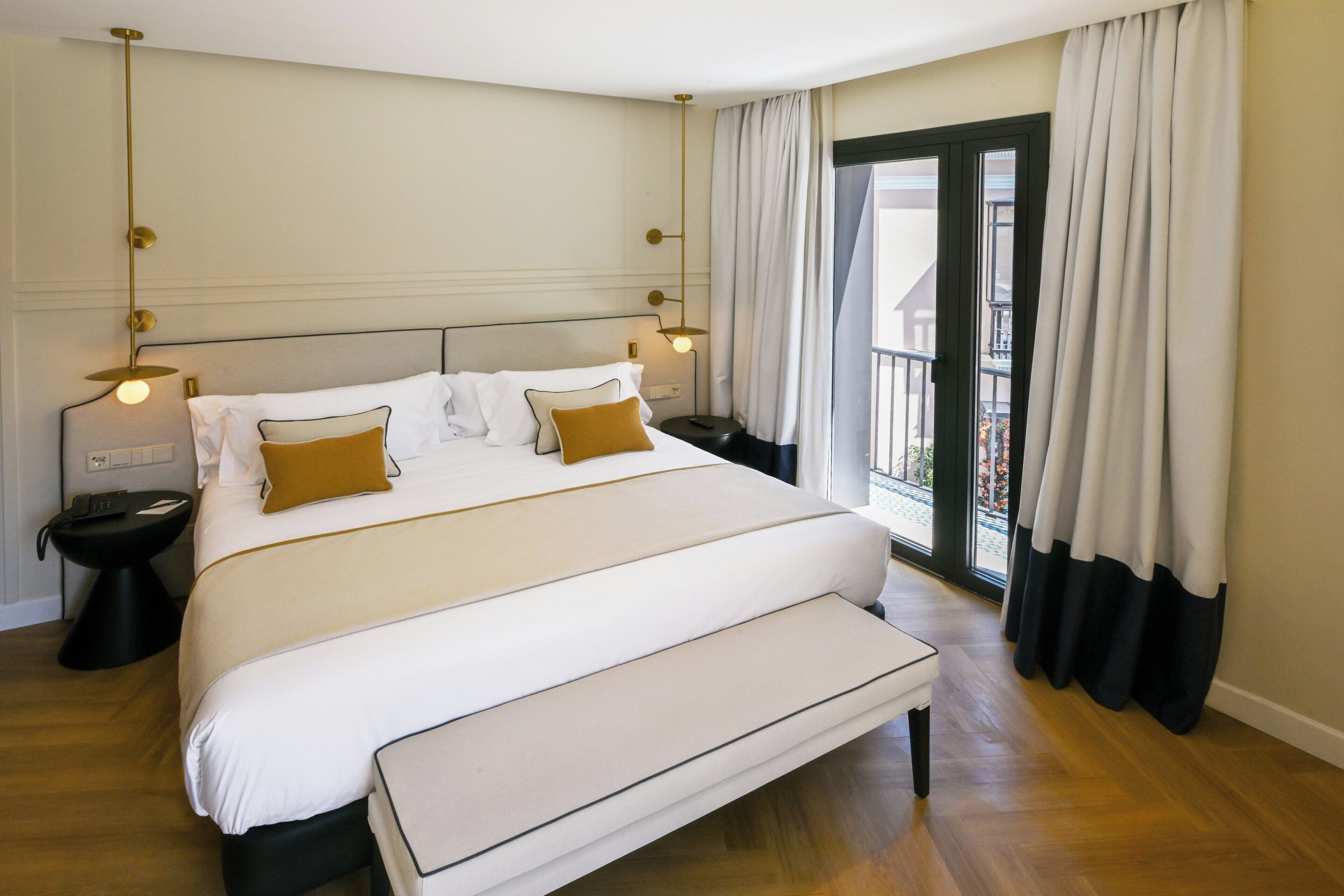 Ein Hotelzimmer mit einem großen Bett, Nachttischen, einer Bank, einem Fenster mit Vorhängen und Balkonblick.
