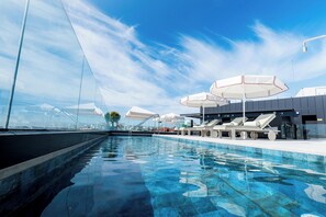 Une piscine avec des chaises longues et des parasols, une barrière en verre et un ciel dégagé.