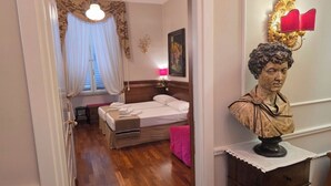 Une chambre d’hôtel avec un plancher en bois, un lit avec du linge de maison blanc, une table de chevet avec une lampe et un buste posé sur une commode.