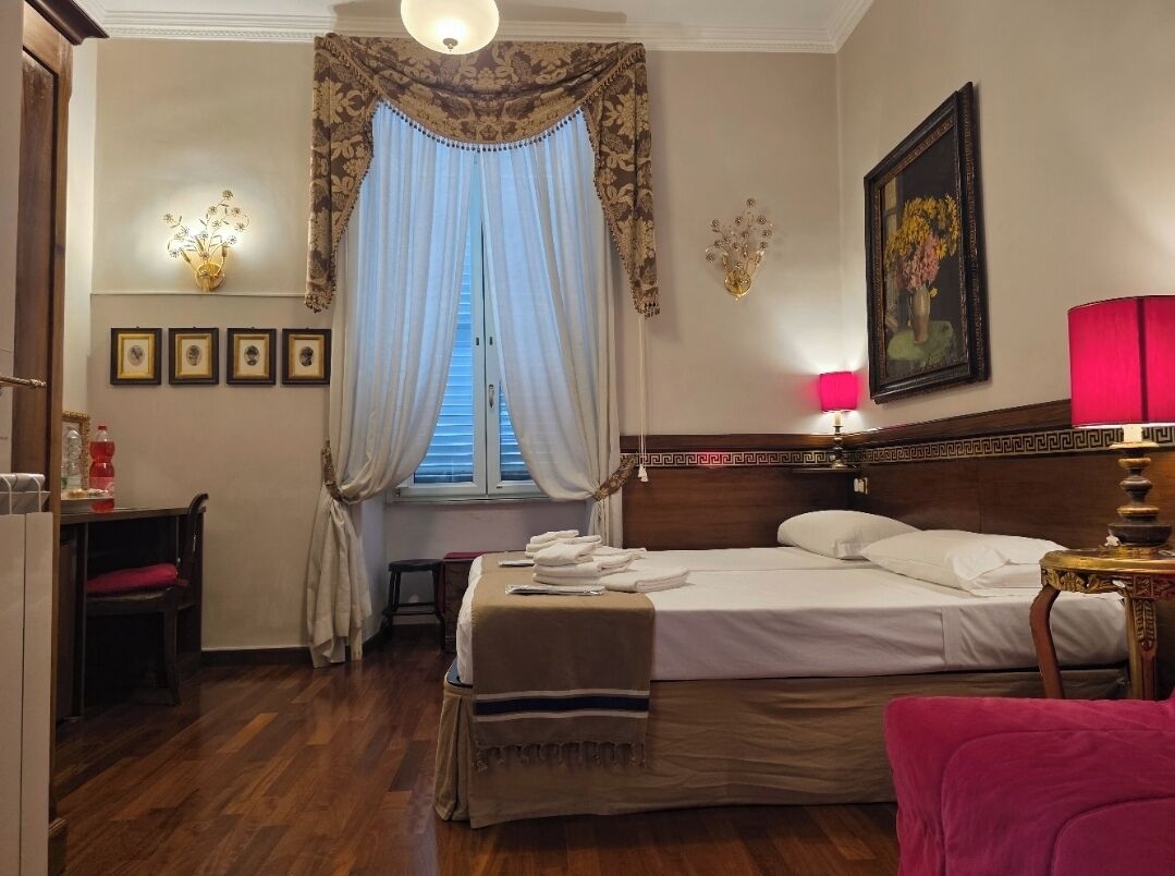 Une chambre d’hôtel comprenant un lit, un bureau, une chaise, une fenêtre avec des rideaux, une lampe fixée au mur et des tableaux encadrés.