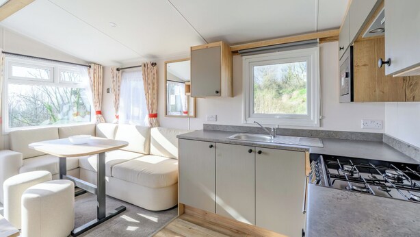 A compact kitchen with a gas stove, a sink, and a window. There is a small dining area with a table and chairs. - Ocean Wave Plus (Pets Welcome) (Woolacombe)