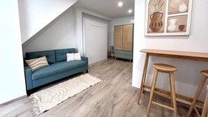 Un soggiorno moderno con un divano color petrolio, un tappeto bianco e un'area pranzo in legno con due sgabelli.