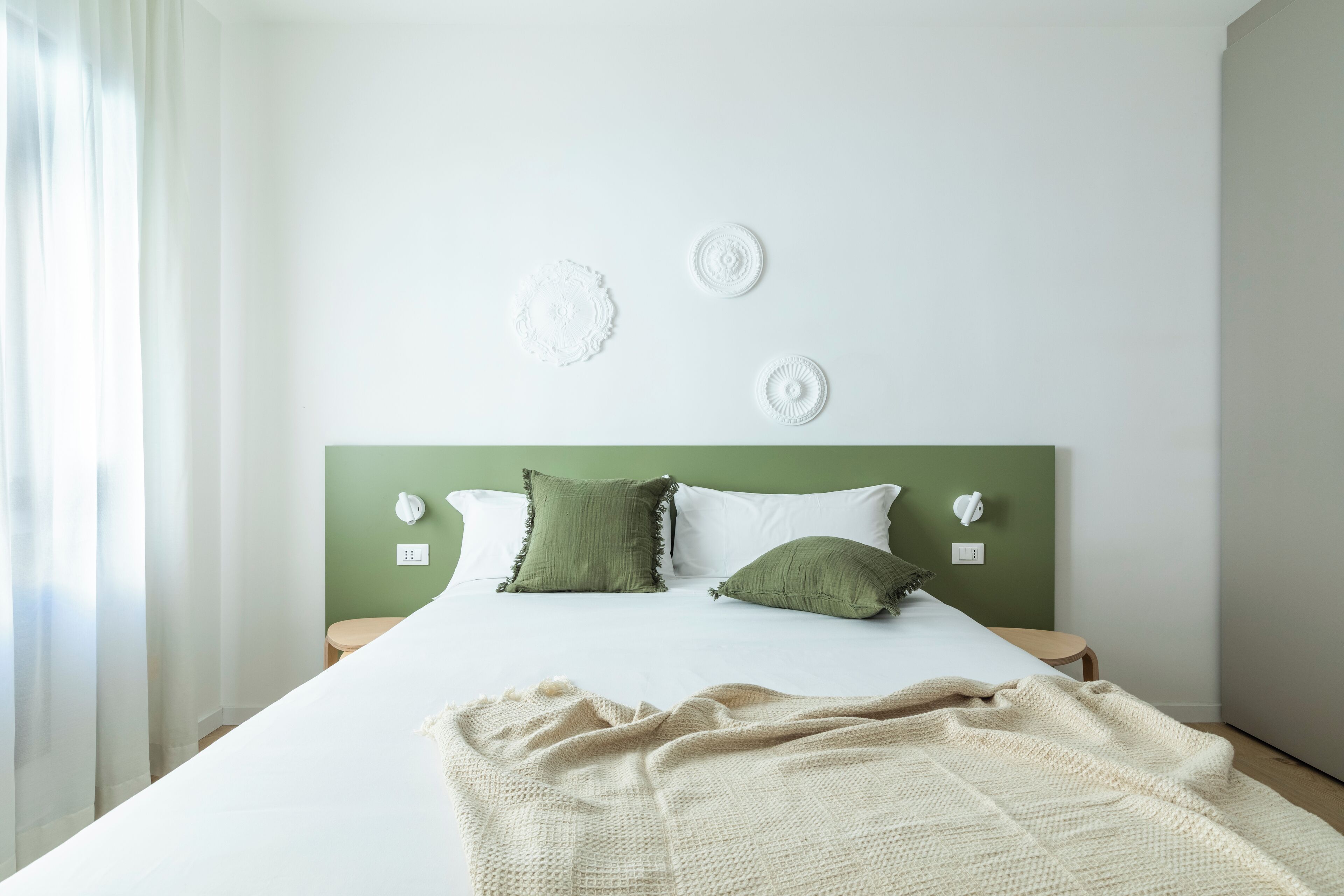 A neatly made bed with a white comforter and two green pillows, a cream-colored knitted blanket, and a green headboard.