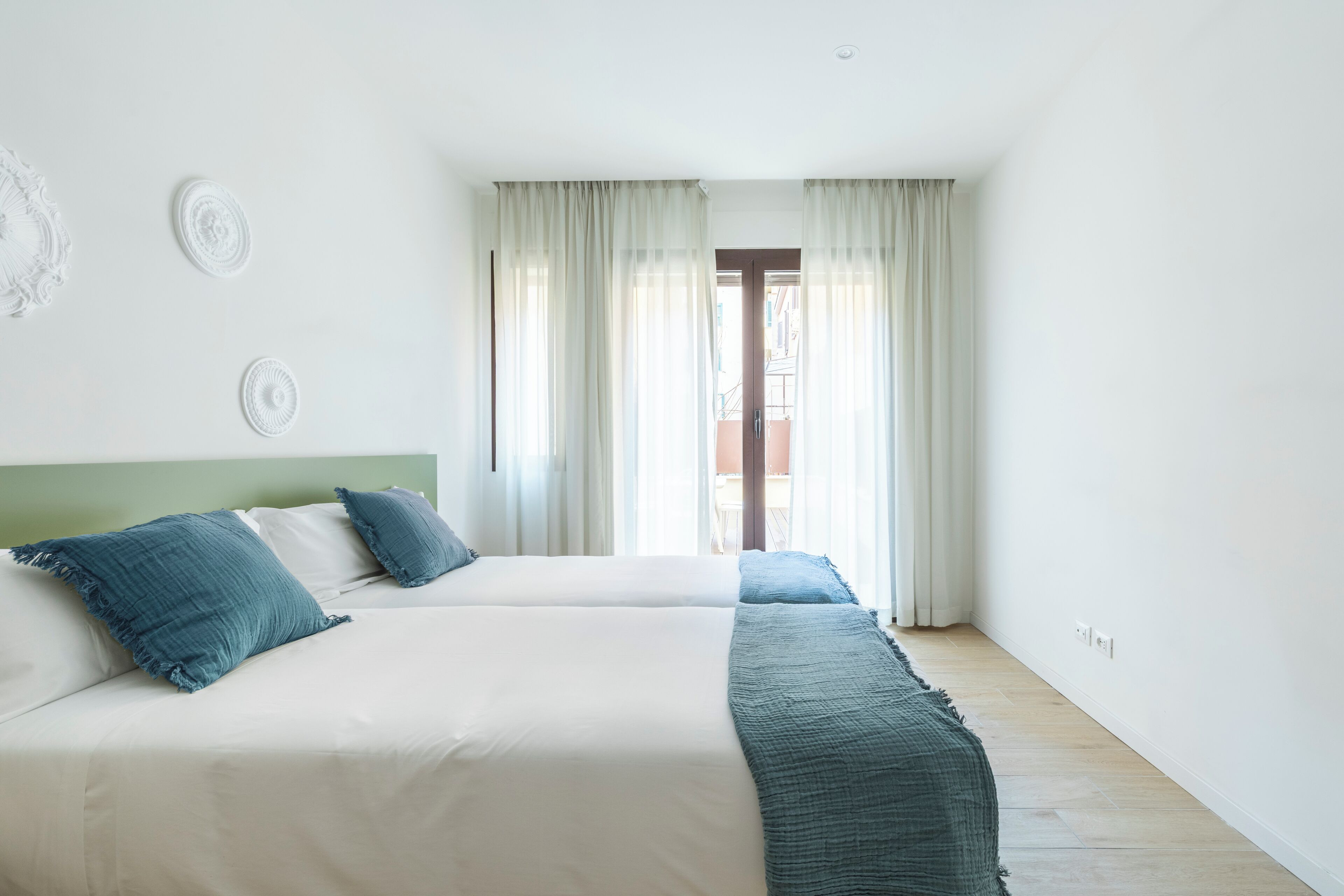 A hotel room with two beds, a green headboard, and a view of the street through sheer curtains.