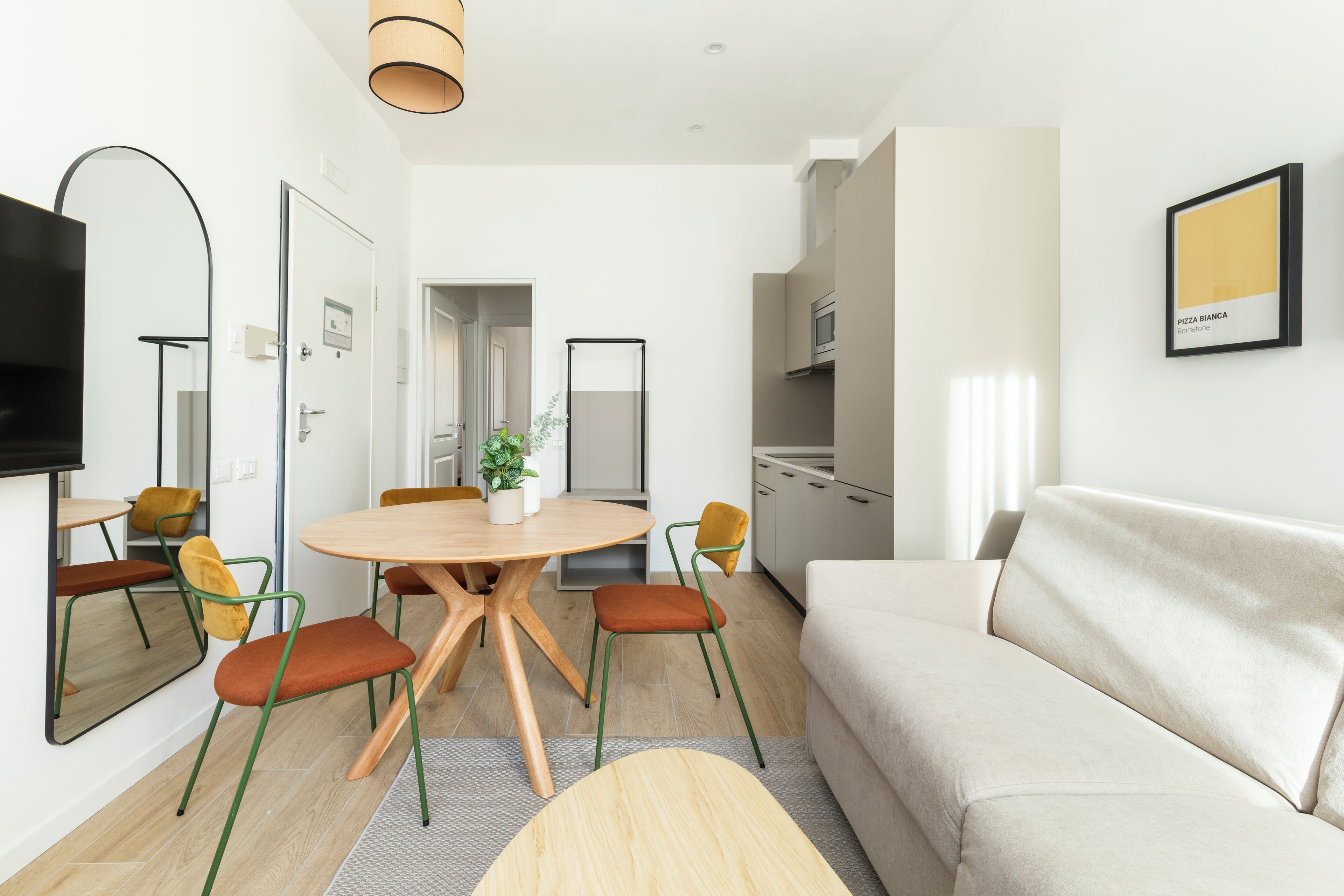 A modern living room with a round wooden table, orange chairs, a beige sofa, and a flat-screen TV mounted on the wall.