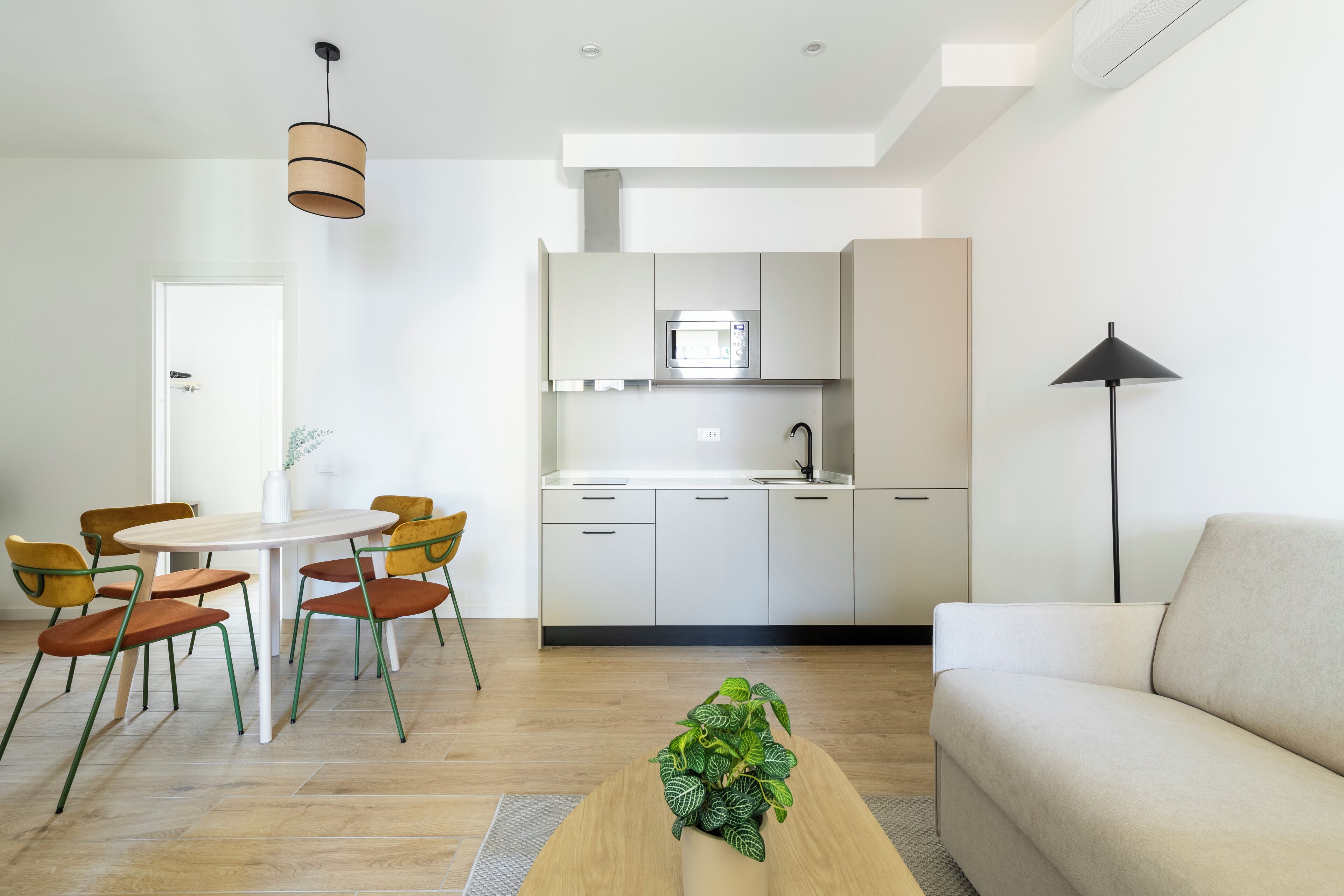 A modern living room with a sofa, a dining table with chairs, a kitchen area with cabinets and a microwave, and a potted plant.