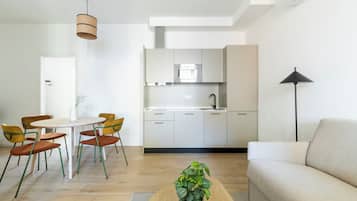 A modern living room with a sofa, a dining table with chairs, a kitchen area with cabinets and a microwave, and a potted plant.