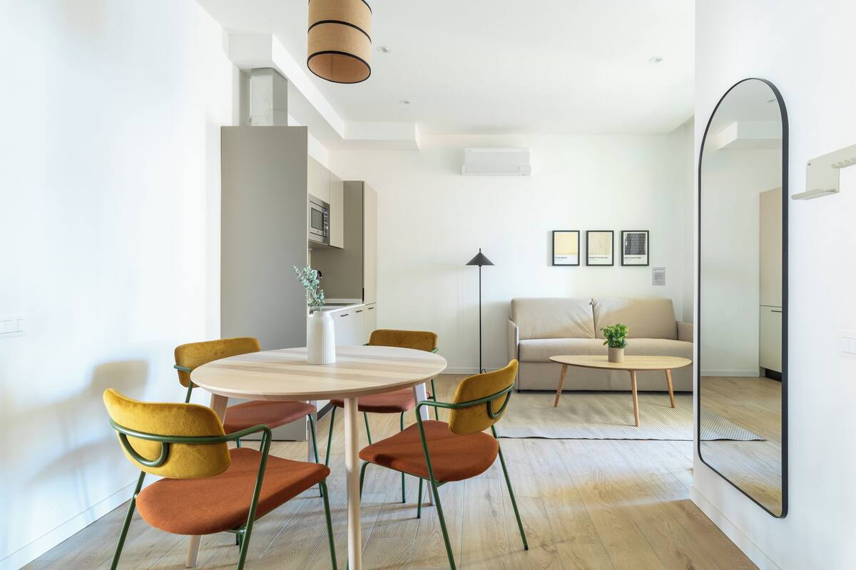 A modern living room with a round dining table, yellow and brown chairs, a beige sofa, and a large mirror.