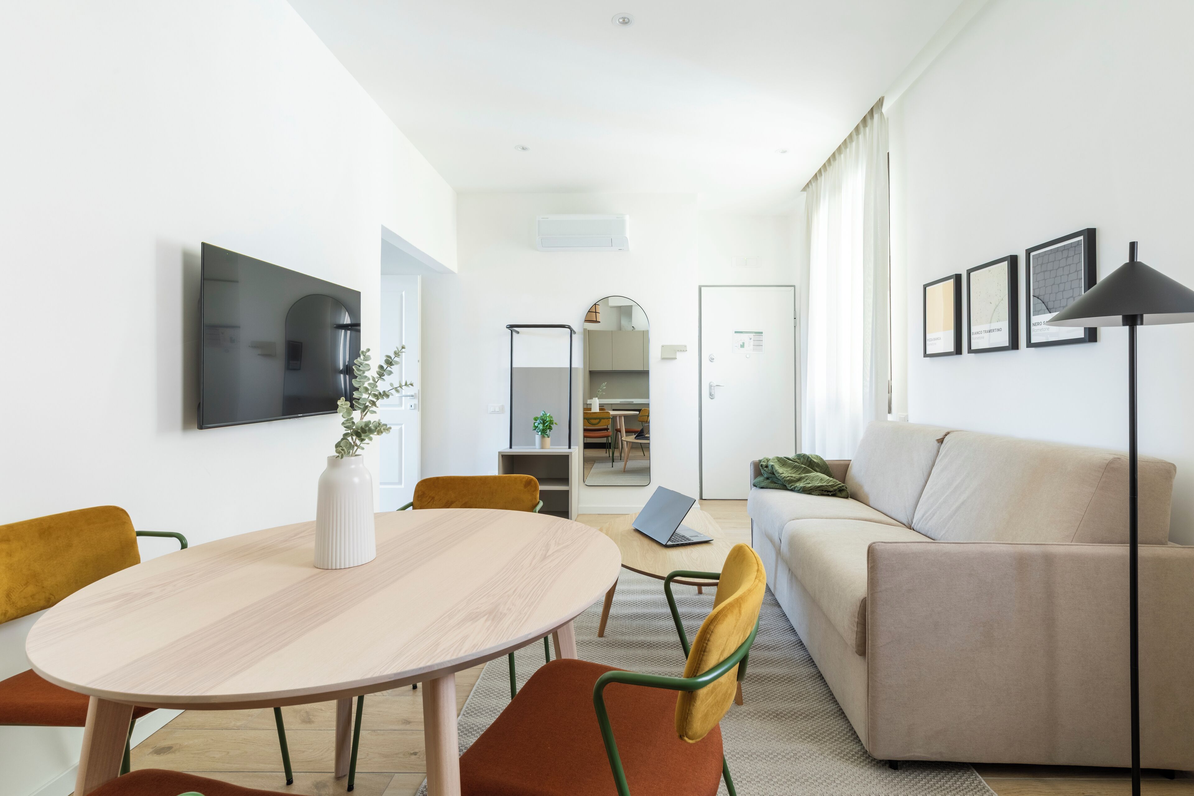 A modern living room with a beige sofa, a round wooden table, and a TV mounted on the wall.