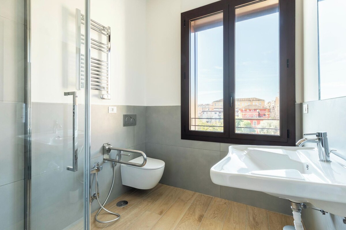 A modern bathroom with a glass shower enclosure, a white toilet, a sink, and a window with a view of buildings.