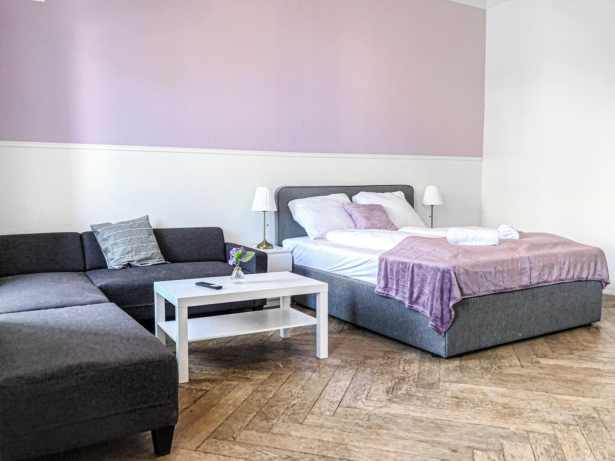 Une chambre d’hôtel moderne avec un canapé gris, une table basse blanche, un lit avec une tête de lit grise et une grande fenêtre.