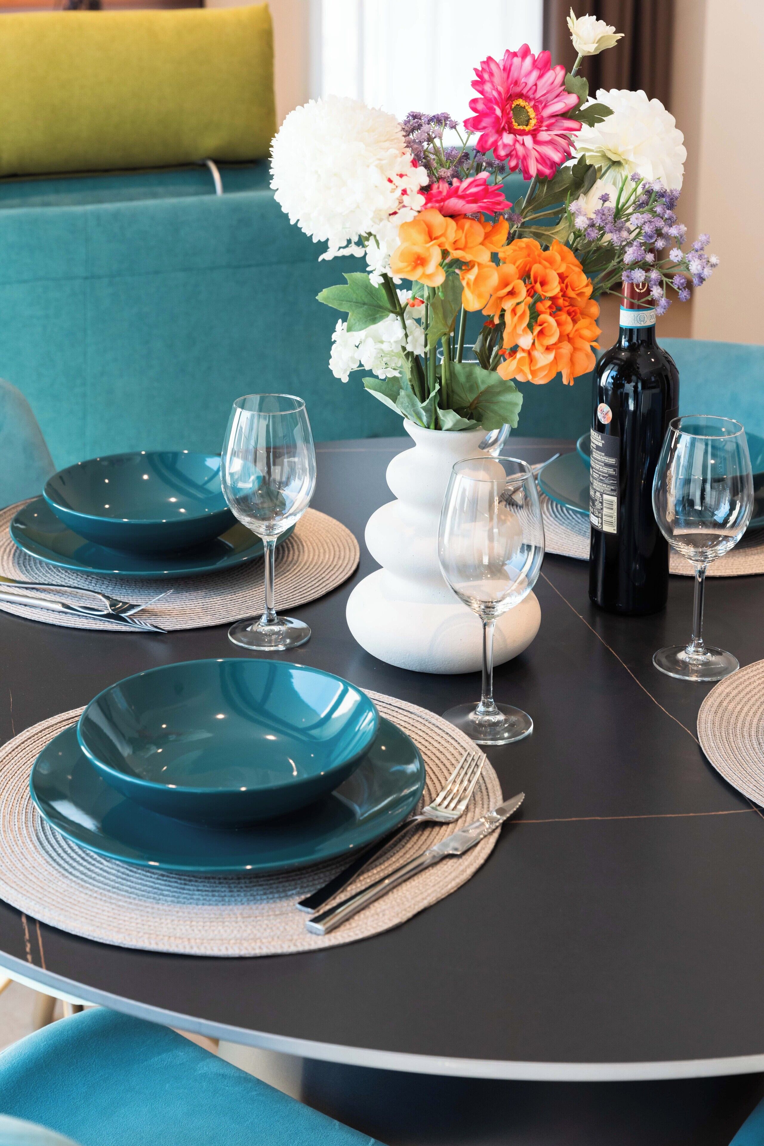Een eettafel met teal borden, wijnglazen en een bloevas.