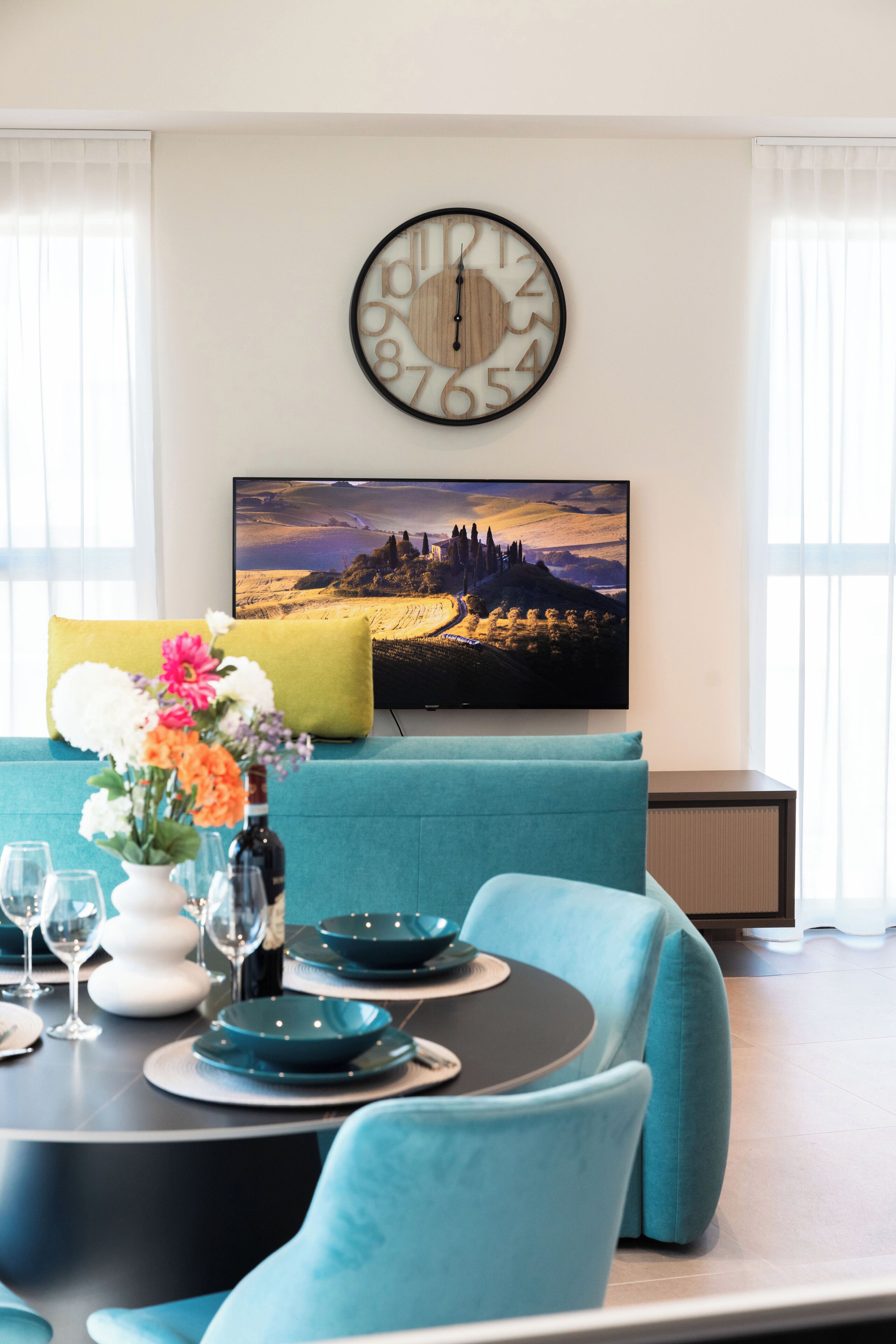 Un comedor con una mesa redonda dispuesta para cuatro personas, un sofá color turquesa, un reloj y un televisor que muestra un paisaje pintoresco.