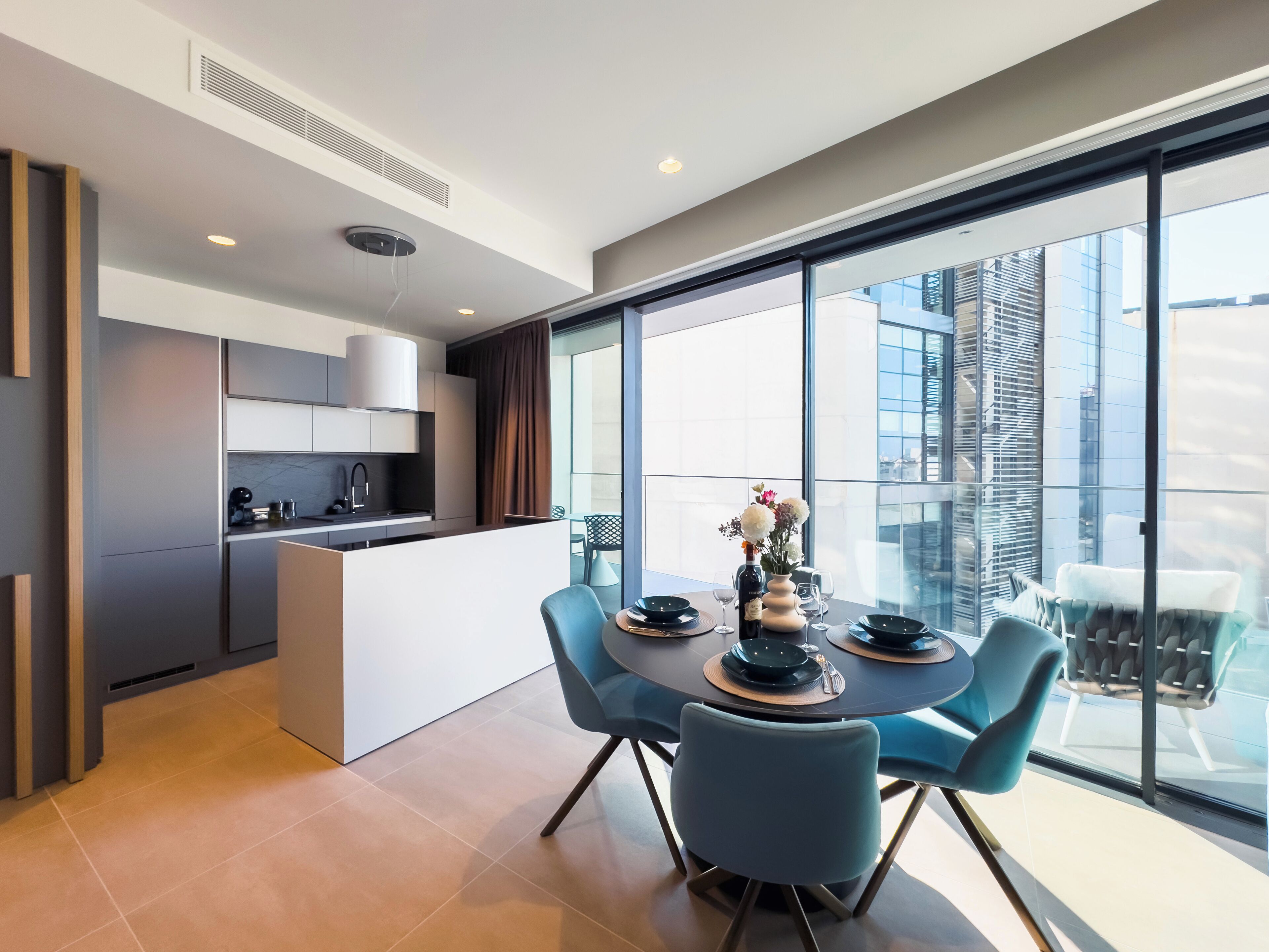A modern kitchen with a central island, a dining area with blue chairs, and large windows offering a city view.