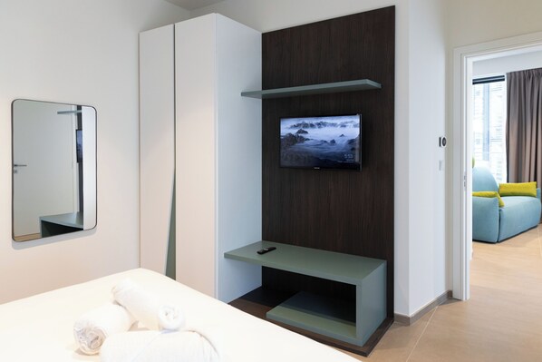 Ein modernes Hotelzimmer mit einem Bett, einem an einer Holzvertäfelung montierten Fernseher, einem Spiegel und Blick auf einen Wohnbereich mit einer Couch.