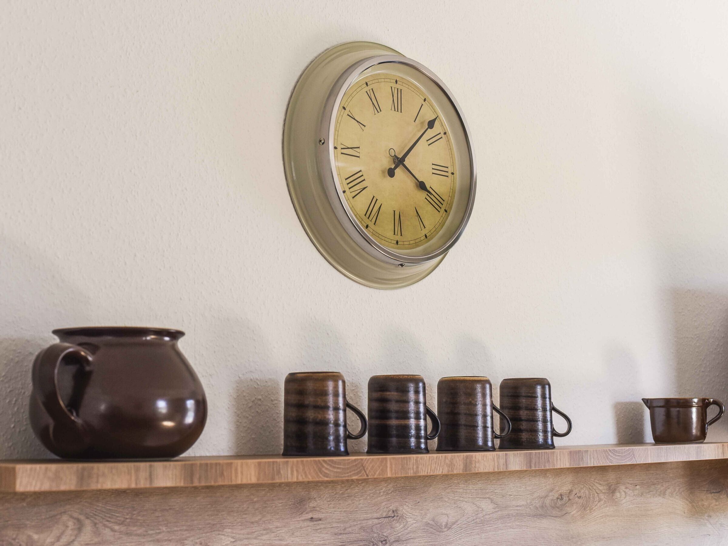 Eine wandmontierte Uhr mit römischen Ziffern, ein Holzregal mit Keramiktassen und eine Keramikkanne.