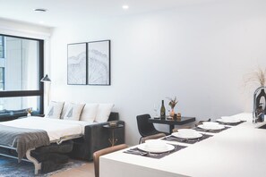 Une chambre moderne avec un lit, une table à manger dressée pour quatre personnes, une petite table d’appoint et une grande fenêtre.
