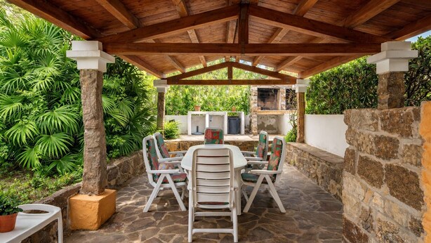 Une terrasse extérieure couverte, dotée d’un sol en pierre, d’une table centrale entourée de chaises et d’un mur en pierre sur l’un des côtés.