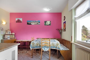 Un coin repas avec un mur rose, des chaises en bois, une table recouverte d’une nappe à motifs floraux et une radio ancienne posée sur une commode.