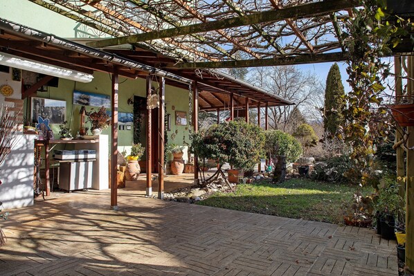Un patio abrité agrémenté d’une pergola, de plantes en pot et d’un chemin en briques menant à l’entrée d’un bâtiment.