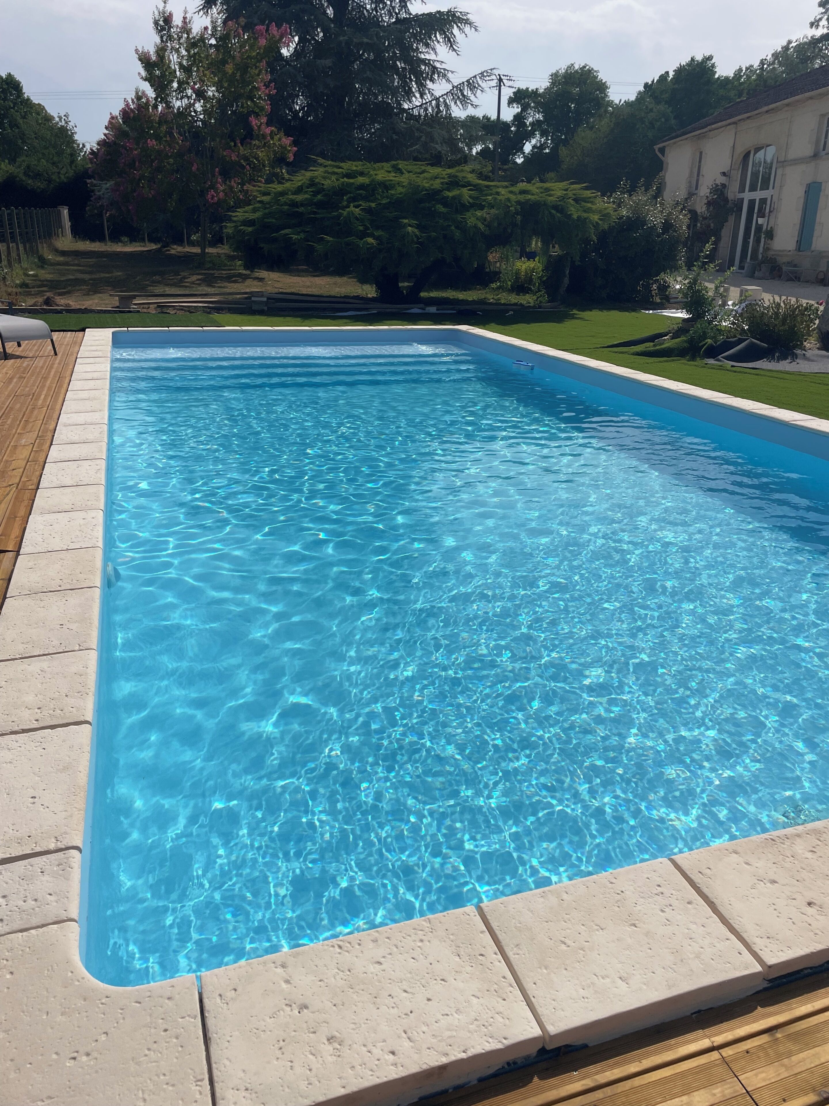 En rektangulær swimmingpool med klart, blåt vand, omgivet af en træterrasse og et græsareal med træer og en bygning i baggrunden.