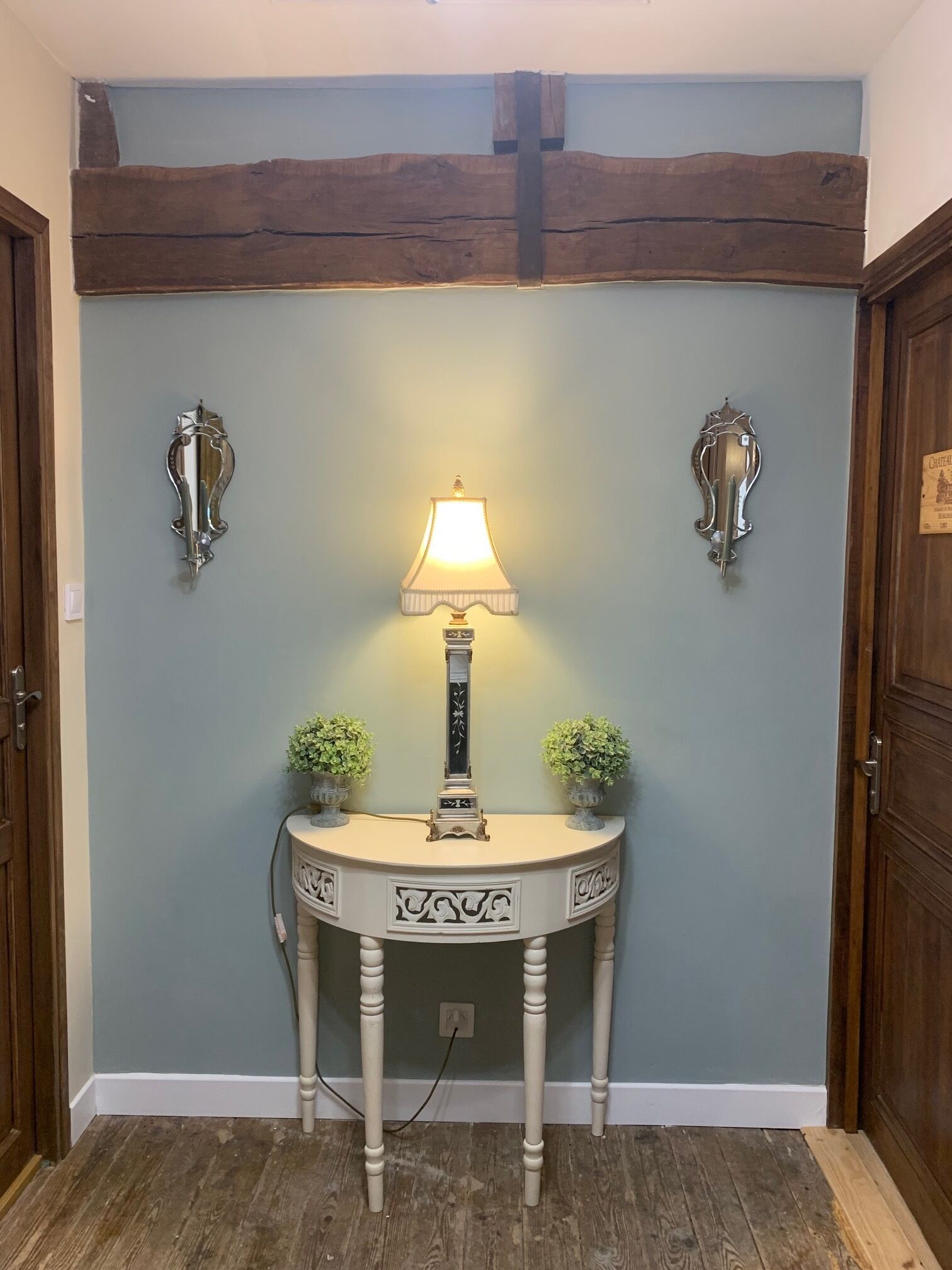 A room with a wooden ceiling, a light blue wall, a white console table with a lamp, two small potted plants, and two wall-mounted sconces.