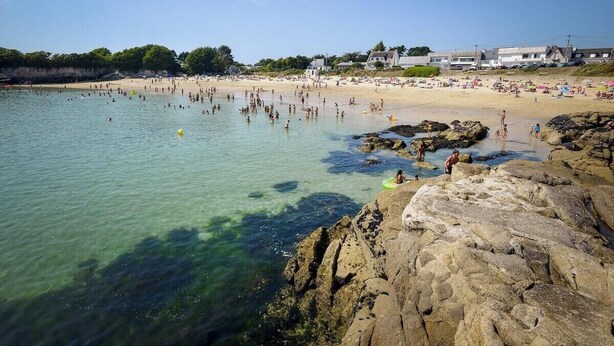 Ein Strand mit klarem Wasser, felsigem Ufer und Badegästen.