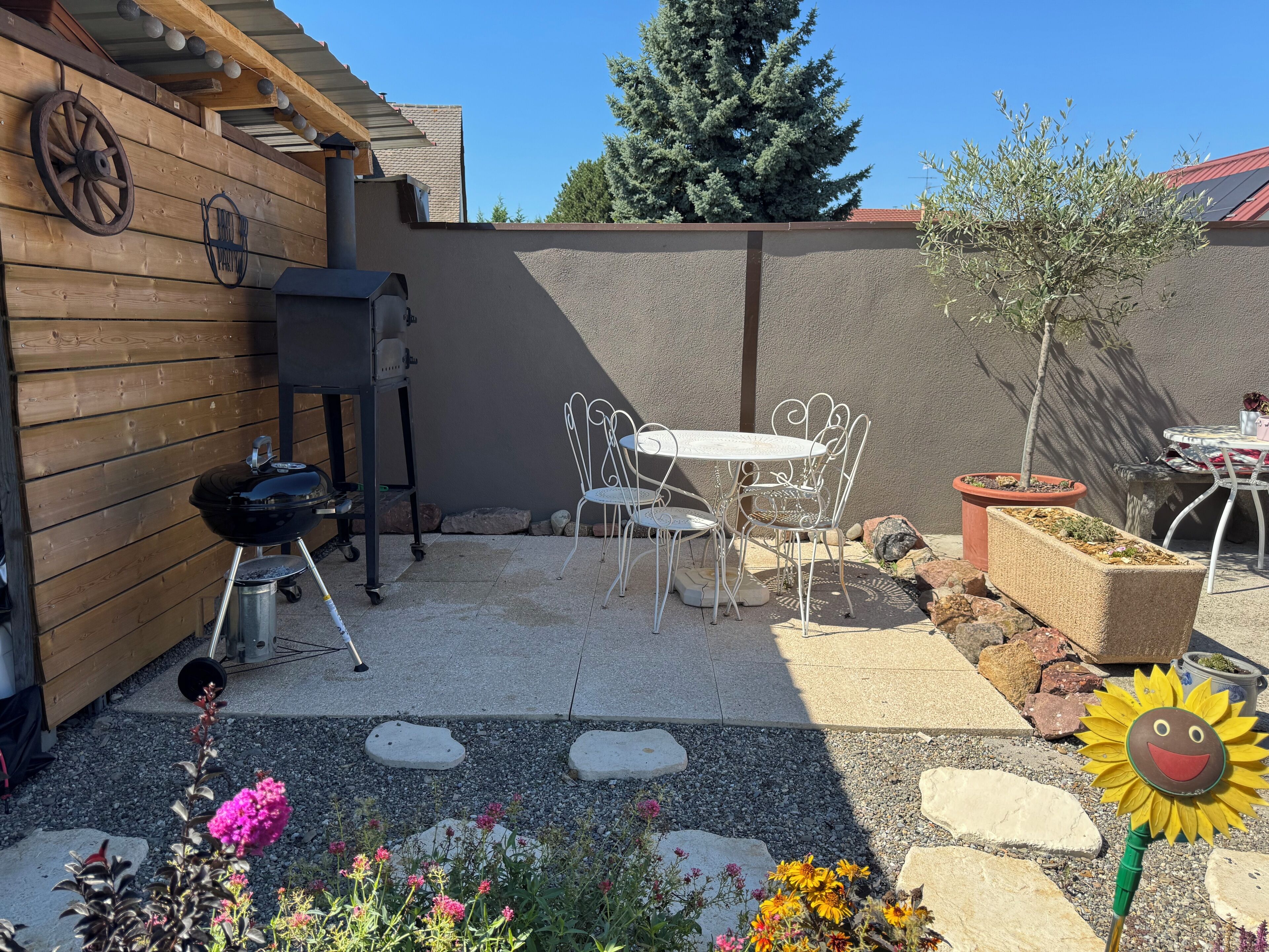 A patio with a grill, white metal chairs, a small table, and potted plants.