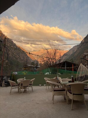 An outdoor seating area with wicker chairs and tables, overlooking a mountainous landscape with a view of a building labeled "Peace Inn." - SolHaus hostel (Kasol)