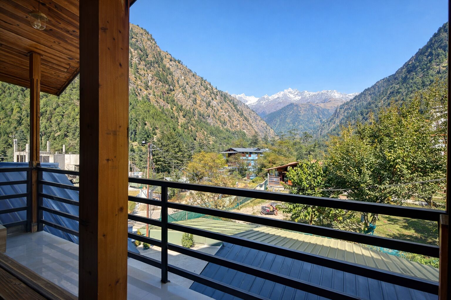 A view from a balcony with a metal railing, overlooking a valley with mountains and a few buildings.