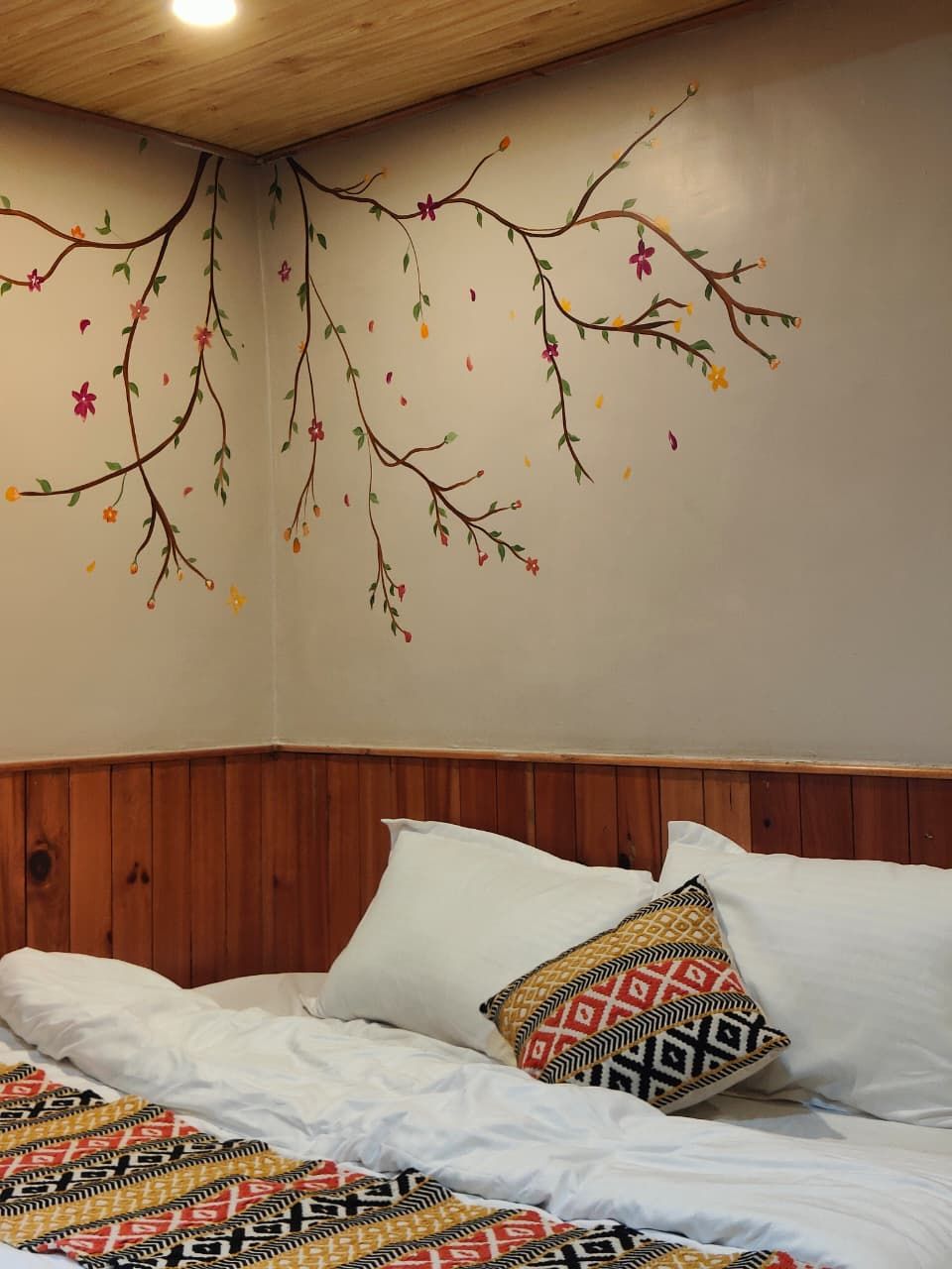 Un dormitorio con cabecera de madera, cama con ropa blanca y un mural en la pared con ramas, hojas y flores.