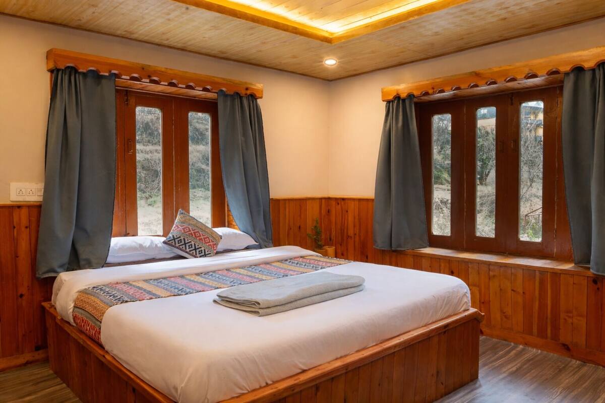 A wooden-framed bedroom with a large bed, two windows with curtains, and a view of trees outside.