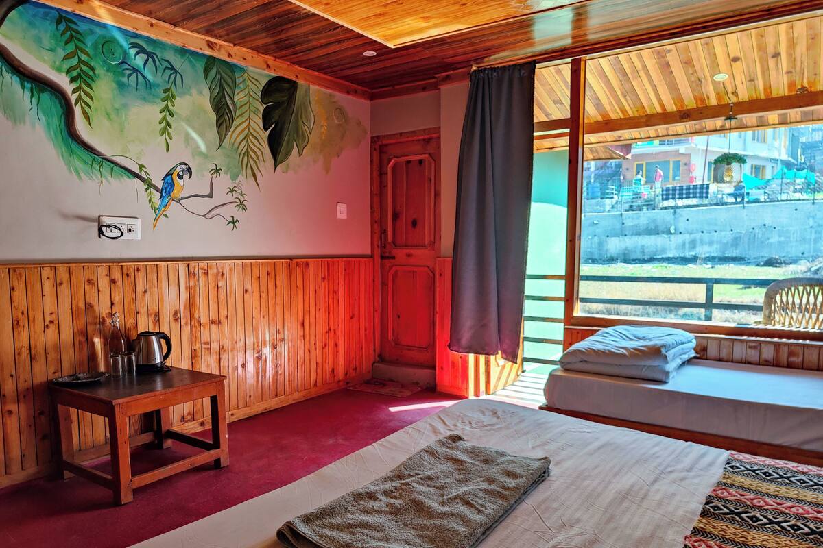 A room with wooden paneling, a bed with a blanket, a small table with a kettle, a wall mural, and a window with a view.