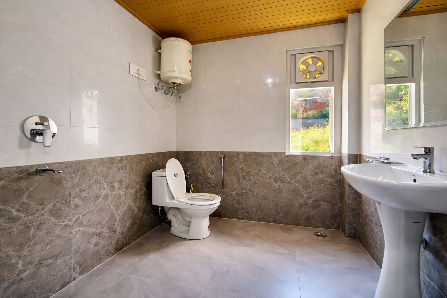 A bathroom with a toilet, a sink, a window with a view, and a fan on the wall.