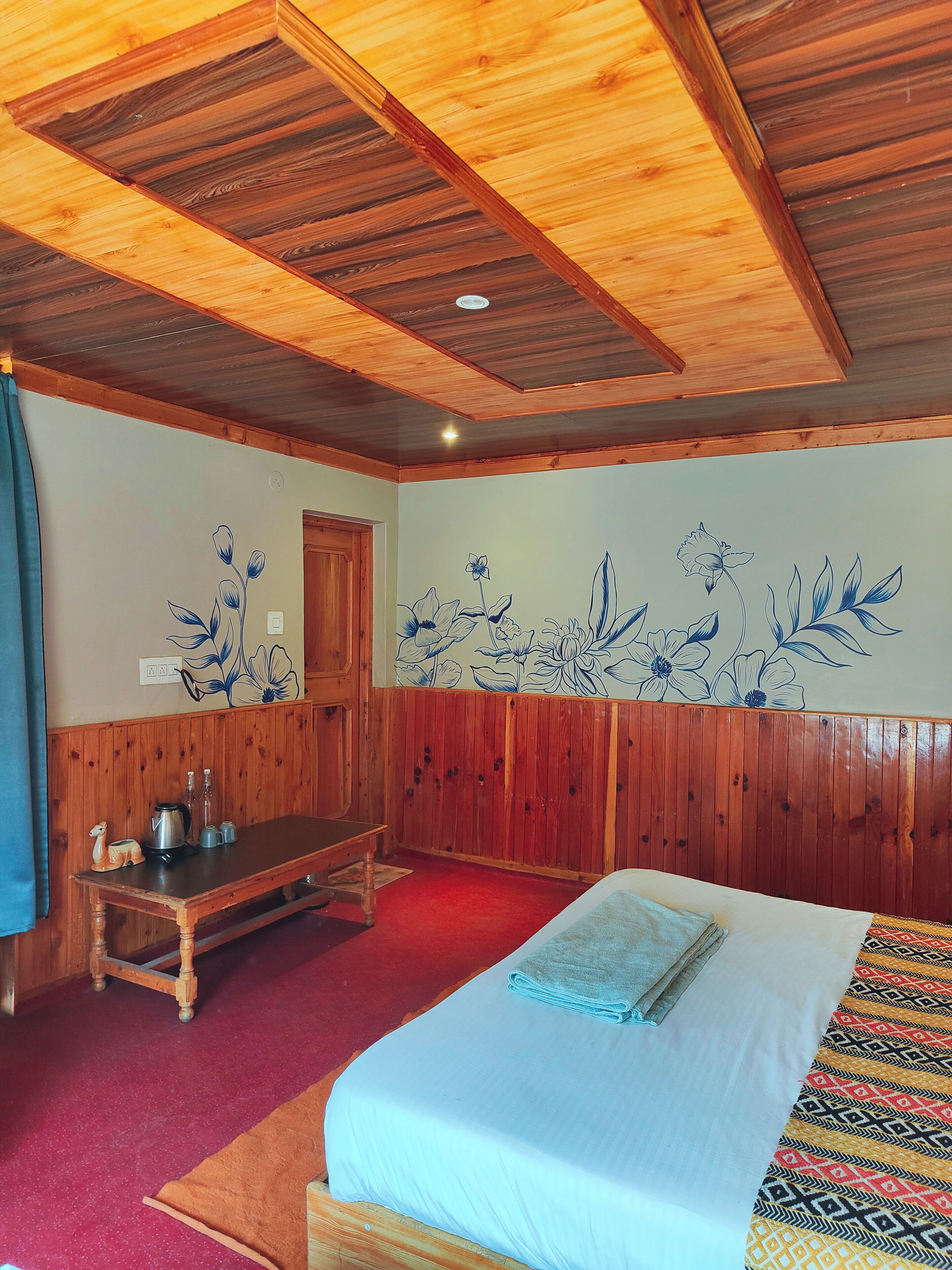 Quarto com painéis de madeira, cama com manta estampada, mesa de madeira com chaleira e parede com arte floral.