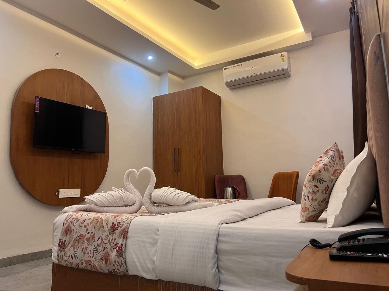 A hotel room with a bed, a TV mounted on a circular wooden panel, a wardrobe, a bedside table with a phone, and an air conditioning unit on the wall.