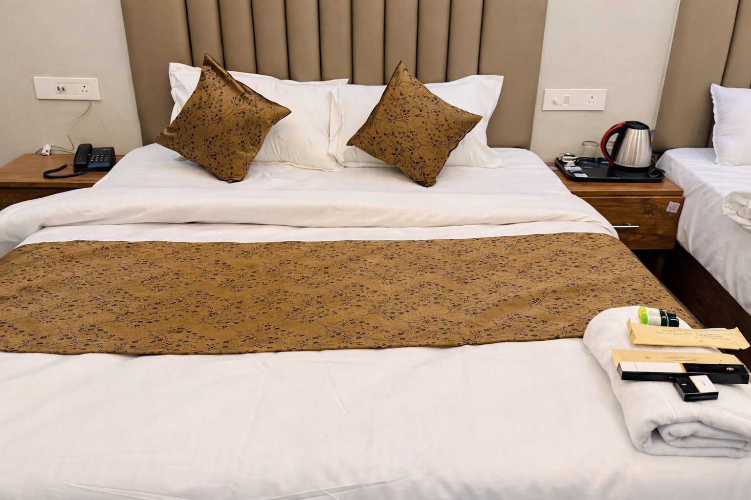 Een net opgemaakt bed met een gebloemd deken, twee decoratieve kussens, een nachtkastje met een telefoon en waterkoker, en een bedtafel met een lamp.