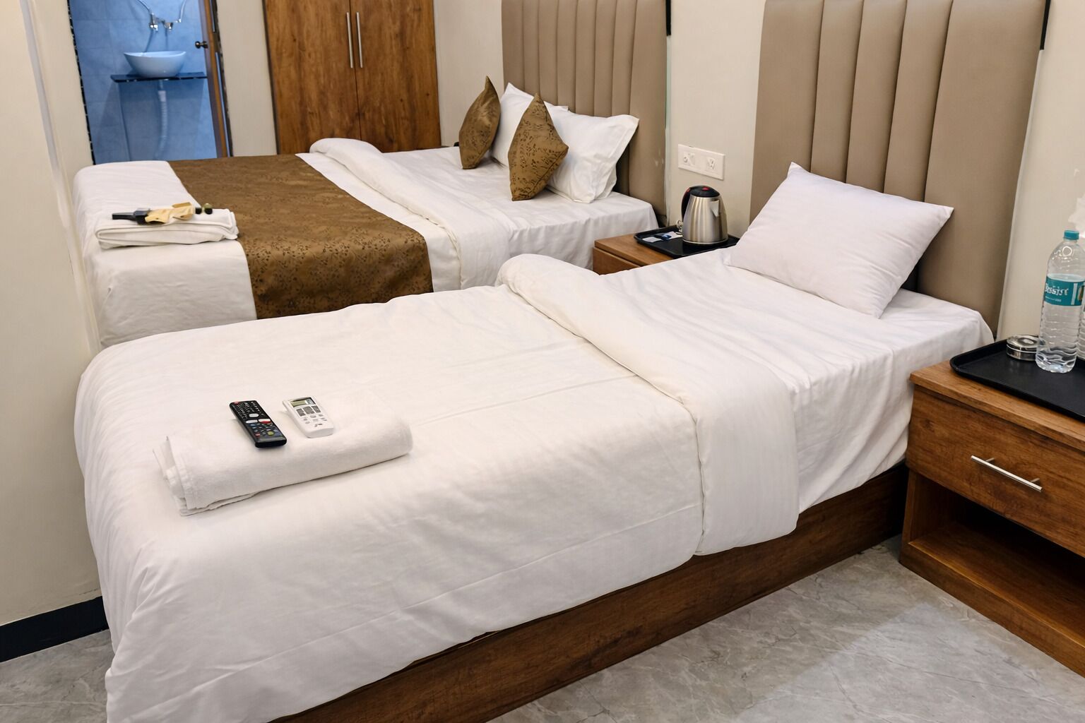 A hotel room with two beds, a wooden wardrobe, a bedside table with a kettle, and a bathroom visible in the background.
