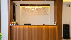 A reception desk with a wooden finish, a modern light fixture above it, and a framed artwork on the wall behind. - Revelon Grand Hotel (Indore)