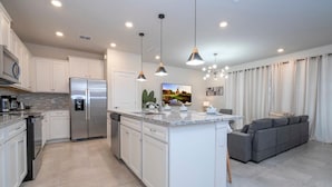 Una cocina moderna con una isla central, electrodomésticos integrados, iluminación colgante y un sofá gris en la zona de estar contigua.