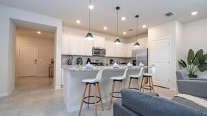 Una cocina moderna con armarios blancos, una isla central con taburetes de bar, iluminación colgante y un sofá gris en la sala de estar.