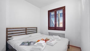 Ein ordentlich bezogenes Bett mit weißen Bettwäsche und Handtüchern, ein Holz-Kopfteil, ein Fenster mit Blick auf Gebäude und ein Heizkörper unter dem Fenster.