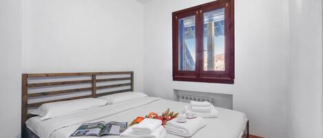 A neatly made bed with white linens and towels, a wooden headboard, a window with a view of buildings, and a radiator under the window.